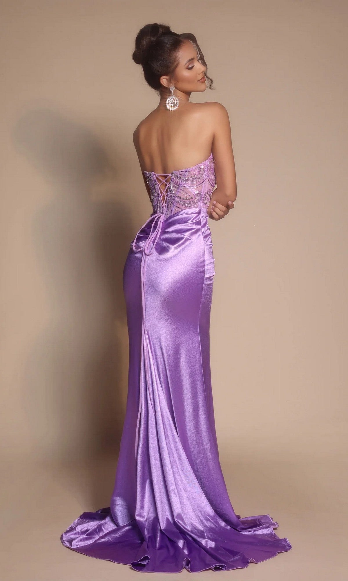 A woman poses with her back to the camera in the Portia and Scarlett XO065 Long Formal Dress, featuring a strapless embellished corset bodice, lace-up back, sequin details, and a lavender satin floor-length skirt against a beige background.